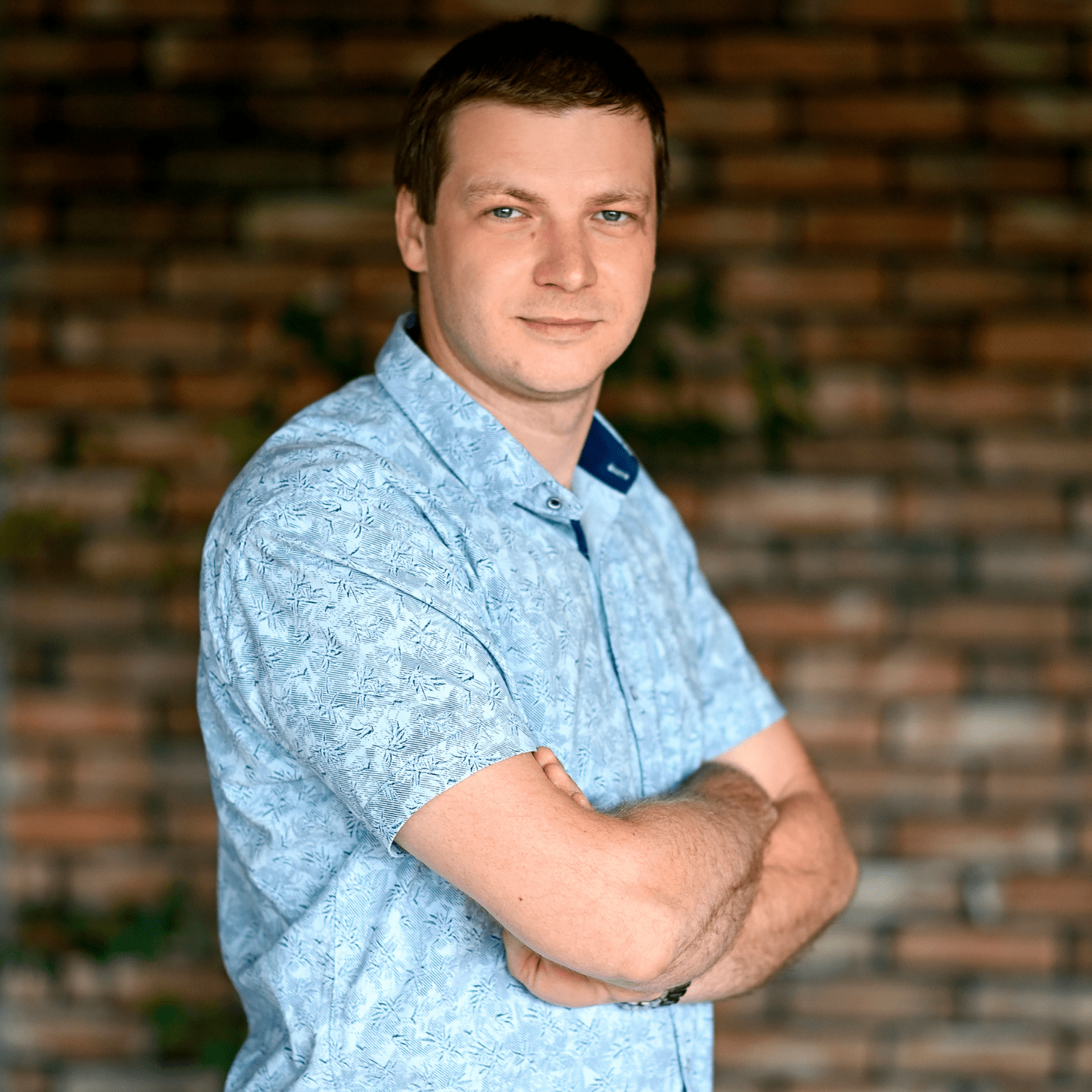 Mężczyzna w niebieskiej koszuli z krótkim rękawem, stojący z założonymi rękami, na tle ceglanej ściany. Idealne ujęcie dla producentów myjni bezdotykowych, podkreślające profesjonalizm i nowoczesność.