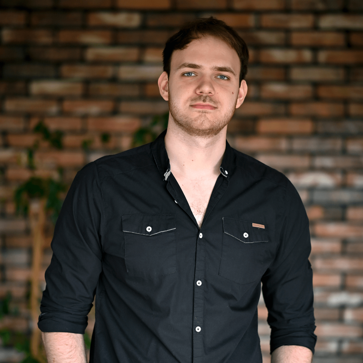 Mężczyzna o jasnych włosach stoi przed ceglaną ścianą, ubrany w czarną koszulę z długim rękawem. Jego postawa i wyraz twarzy emanują pewnością siebie. W tle widoczne są rośliny, co nadaje zdjęciu świeżości. Idealny obraz do promocji myjni bezdotykowych i ich producentów.