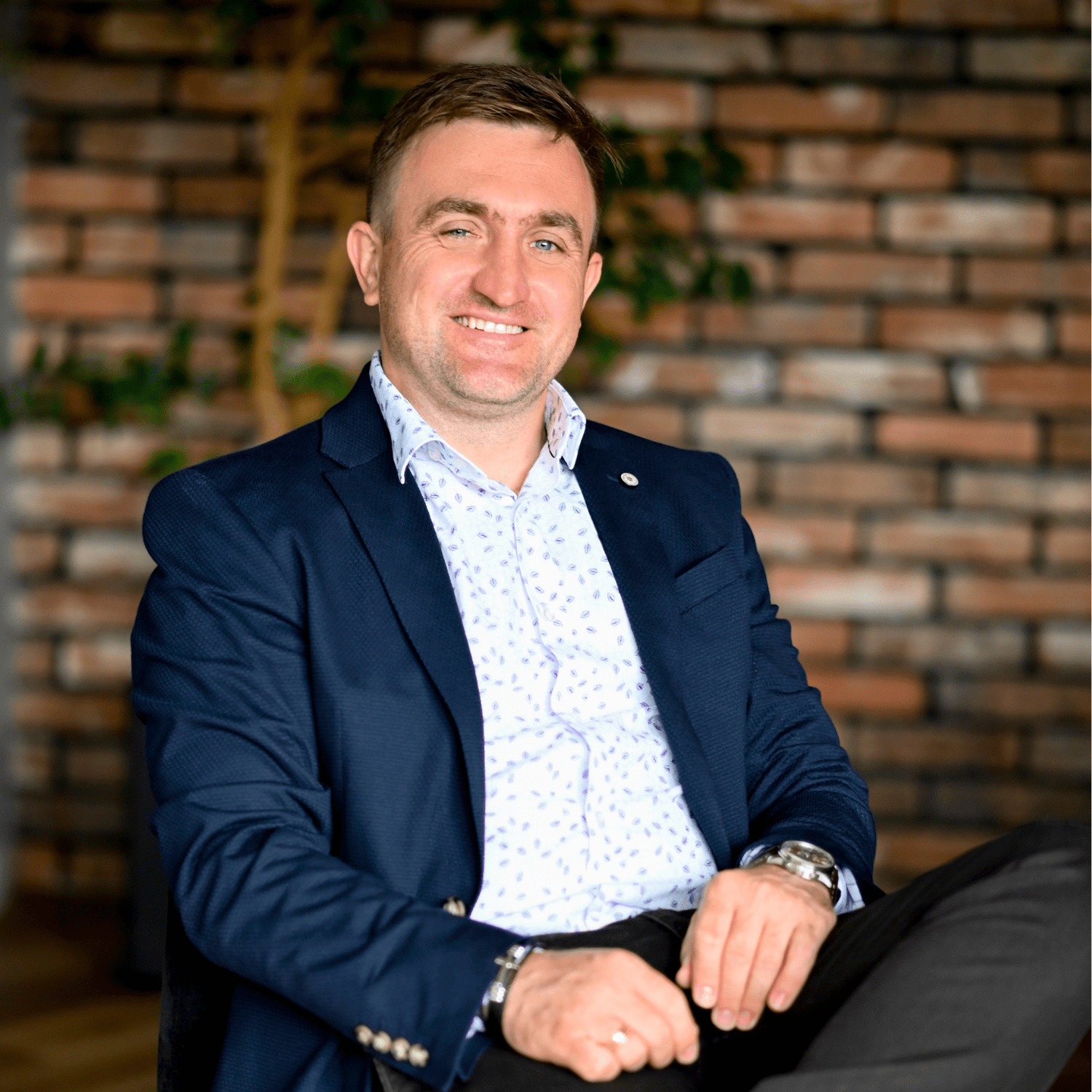 Mężczyzna w granatowym garniturze siedzi na kanapie w nowoczesnym biurze, uśmiechając się do kamery. W tle widoczna jest ceglana ściana i roślinność, co tworzy przyjemną atmosferę. Producent myjni bezdotykowych prezentuje profesjonalny wizerunek.