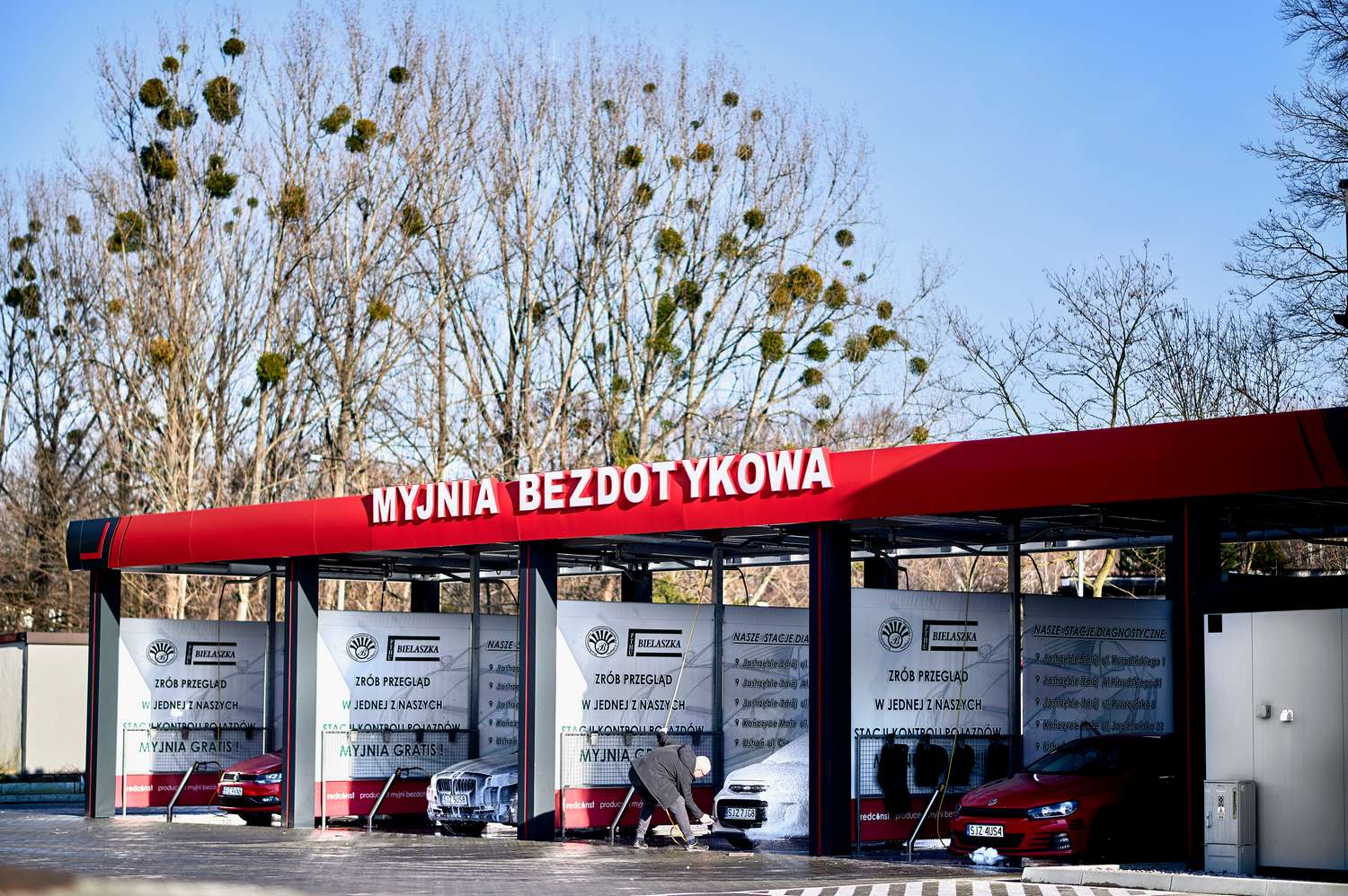 Zewnętrzny widok myjni bezdotykowej z czerwonym dachem i wieloma stanowiskami do mycia samochodów. W tle widać drzewa, a w pierwszym planie mężczyzna czyści pojazd. Na banerach widoczne informacje o usługach i promocjach. Idealne dla miłośników motoryzacji w poszukiwaniu sprawdzonego producenta myjni bezdotykowej.
