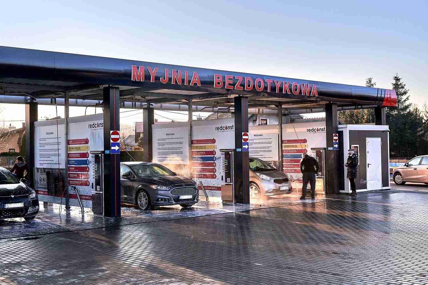 Myjnia bezdotykowa z kilkoma pojazdami, ludźmi korzystającymi z usług oraz ładnym, błyszczącym wykończeniem podłogi. Dobre warunki do mycia samochodów w nowoczesnym otoczeniu.