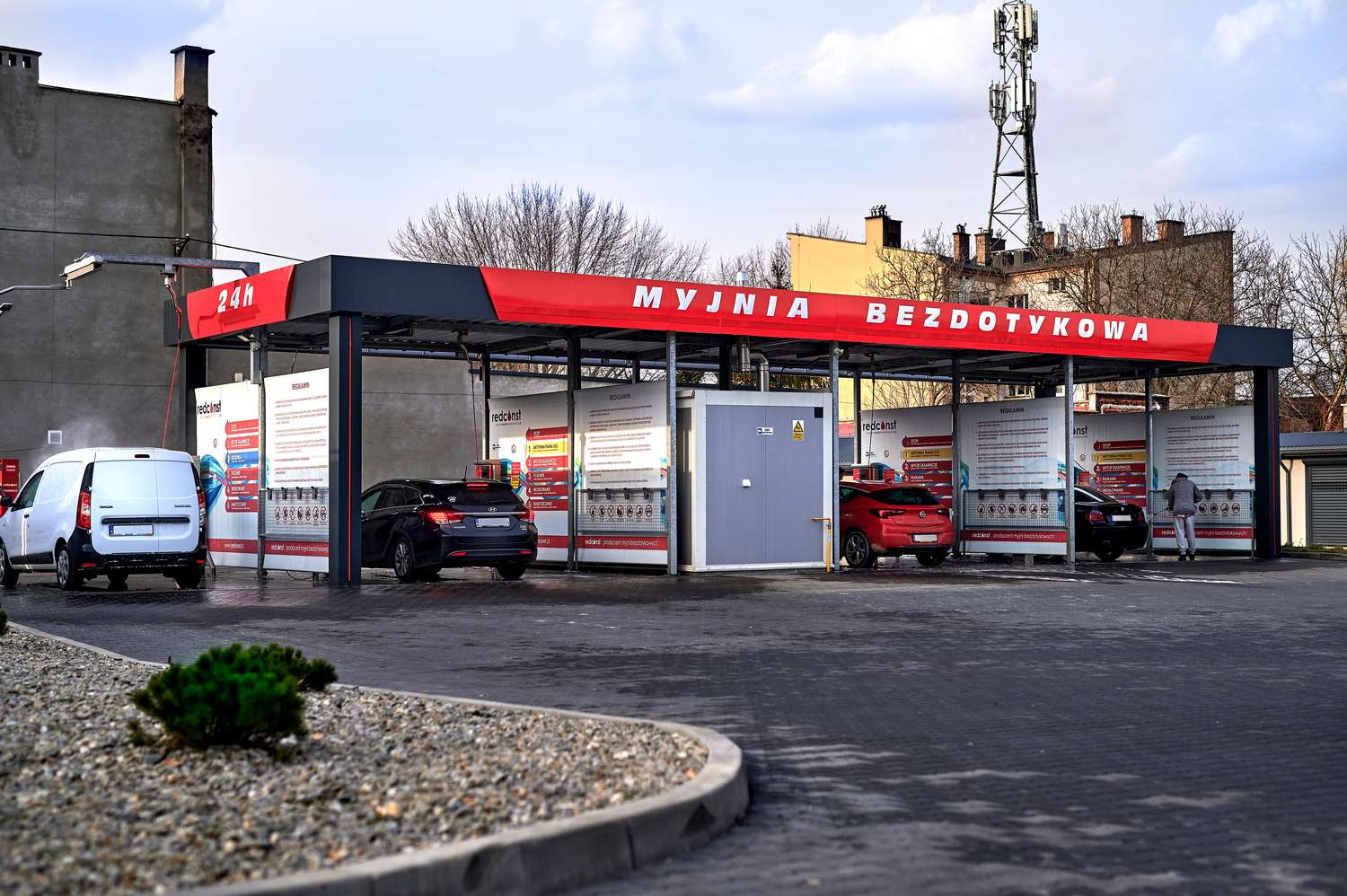 Myjnia bezdotykowa z czerwonym szyldem, oferująca całodobowe usługi mycia samochodów. W tle widać zaparkowane pojazdy oraz efektywne stanowiska myjni. Idealne dla kierowców poszukujących nowoczesnych rozwiązań.