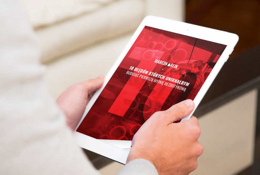 Osoba trzymająca tablet, na którym widoczna jest okładka książki "10 błędów, których uniknęłyś, budując pierwszą myjnię bezdotykową" autorstwa Marcina Orlika. Tło okładki w kolorze czerwonym z graficznymi elementami.