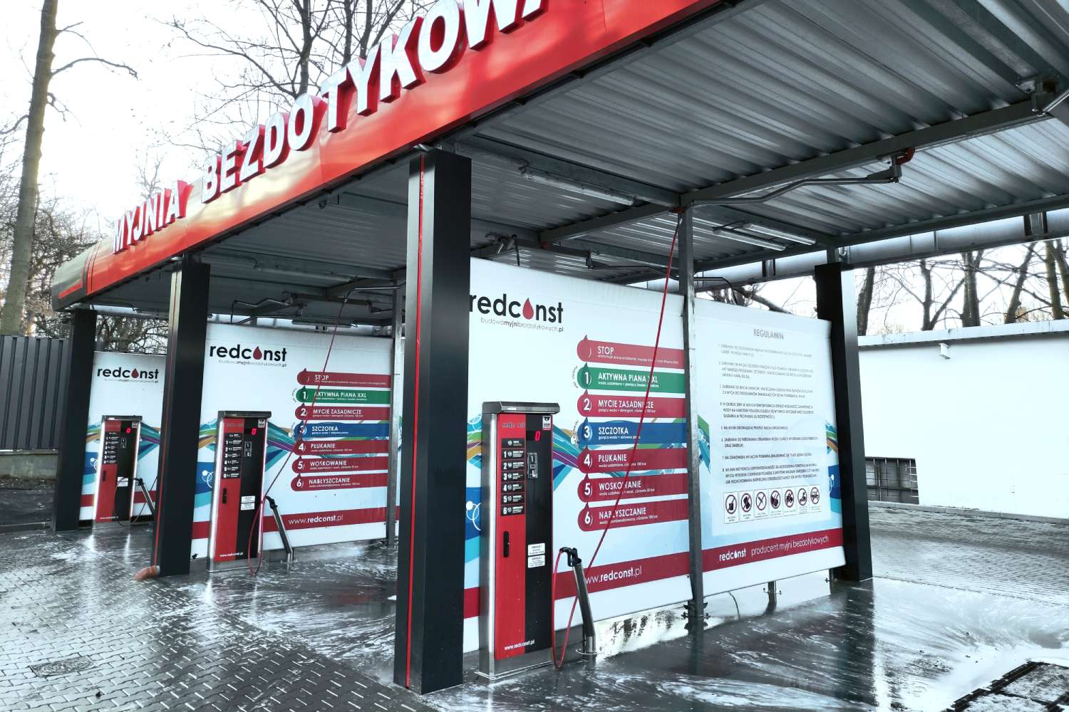 Widok na myjnię bezdotykową z czerwonym szyldem "myjnia bezdotykowa" oraz instrukcjami mycia na ścianie. Widać kilka stanowisk myjących z automatami i wężami. Teren myjni jest mokry, co sugeruje aktywne użytkowanie. To idealne miejsce dla entuzjastów dbania o czystość pojazdów.