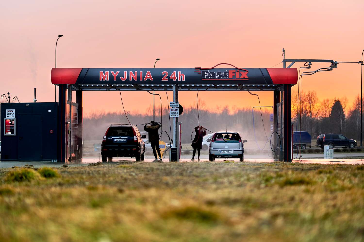 Obraz przedstawia myjnię bezdotykową FastFix działającą 24 godziny, z klientami myjącymi swoje samochody w zimnym powietrzu na tle zachodzącego słońca. Na pierwszym planie widać zadbaną przestrzeń oraz sprzęt do mycia.