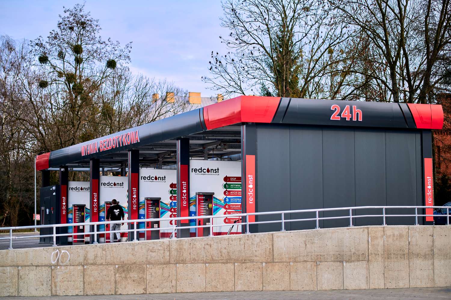 Myjnia bezdotykowa otwarta 24h, oferująca nowoczesne stacje do samodzielnego mycia pojazdów. Na stalowej konstrukcji umieszczono kolorowe urządzenia do obsługi klientów, a w tle widoczne drzewa.