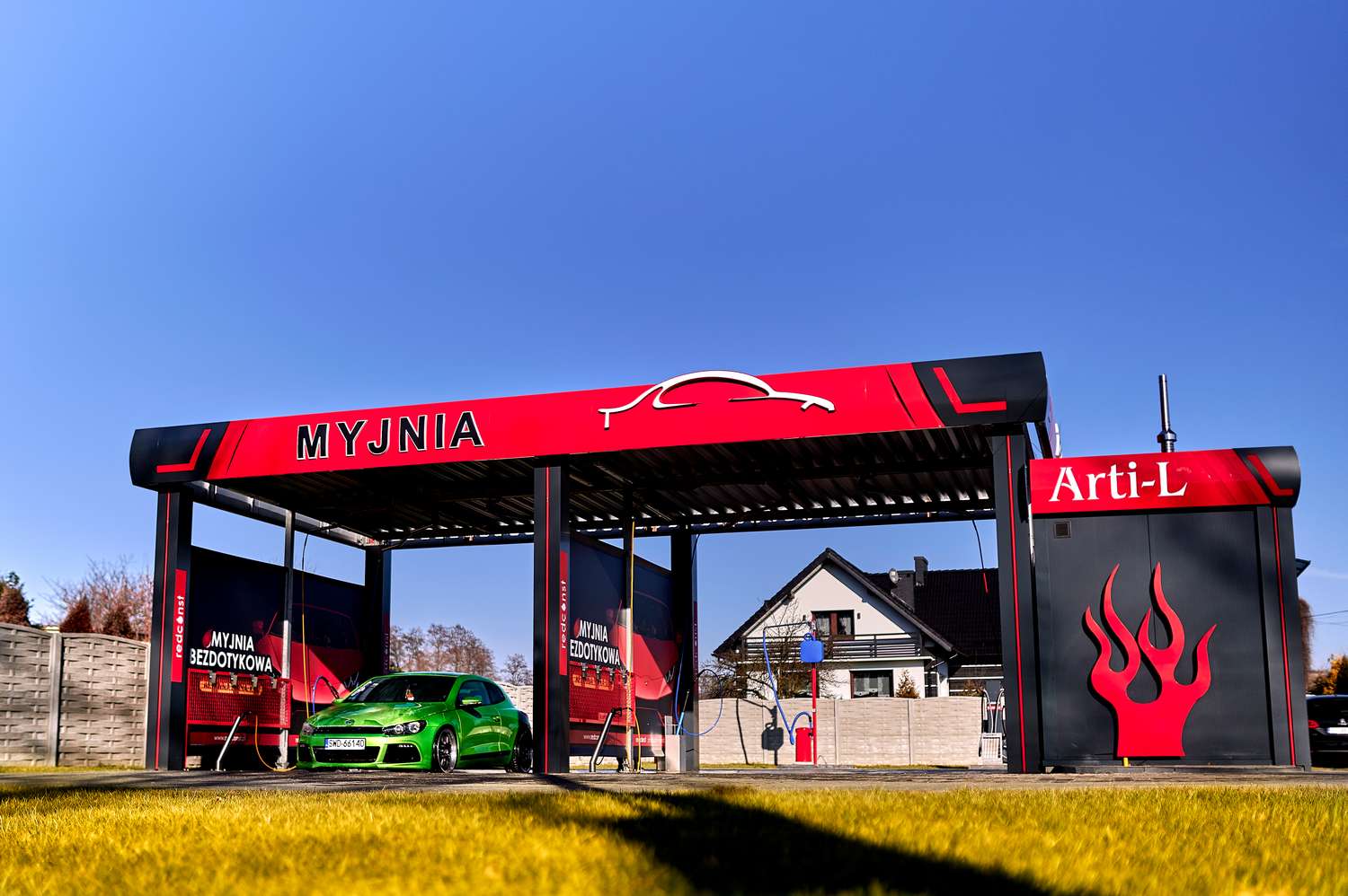 Stacja myjni bezdotykowej z jasnym, zielonym samochodem na pierwszym planie. W tle widoczny dom jednorodzinny oraz błękitne niebo. Z przodu dużą czerwono-czarną tablicą z napisem "MYJNIA" oraz logo producenta Arti-L, wskazującym na nowoczesne usługi czyszczenia pojazdów.