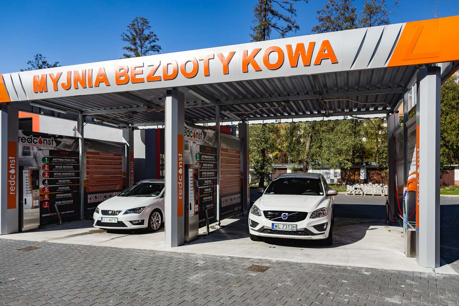 Widok nowoczesnej myjni bezdotykowej z dwoma samochodami pod zadaszeniem. W tle znajduje się wyposażenie, instrukcje oraz kolorowe oznaczenia dla klientów. Użyteczne miejsce dla właścicieli pojazdów poszukujących wygodnych usług myjni bezdotykowej.