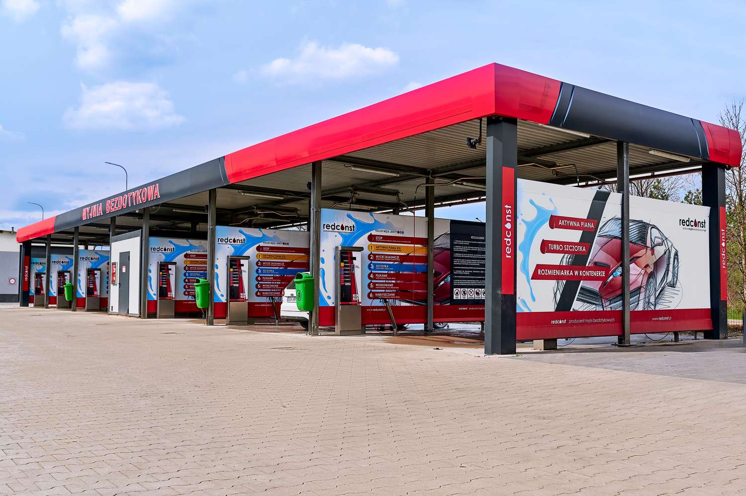 Nowoczesna myjnia bezdotykowa z kilkoma stanowiskami do mycia samochodów, z czytelnymi instrukcjami i ekologicznymi pojemnikami na odpady. W tle widoczna clear blue sky, co tworzy przyjemną atmosferę dla klientów. Myjnie bezdotykowe oferują szybkie i skuteczne czyszczenie pojazdów.