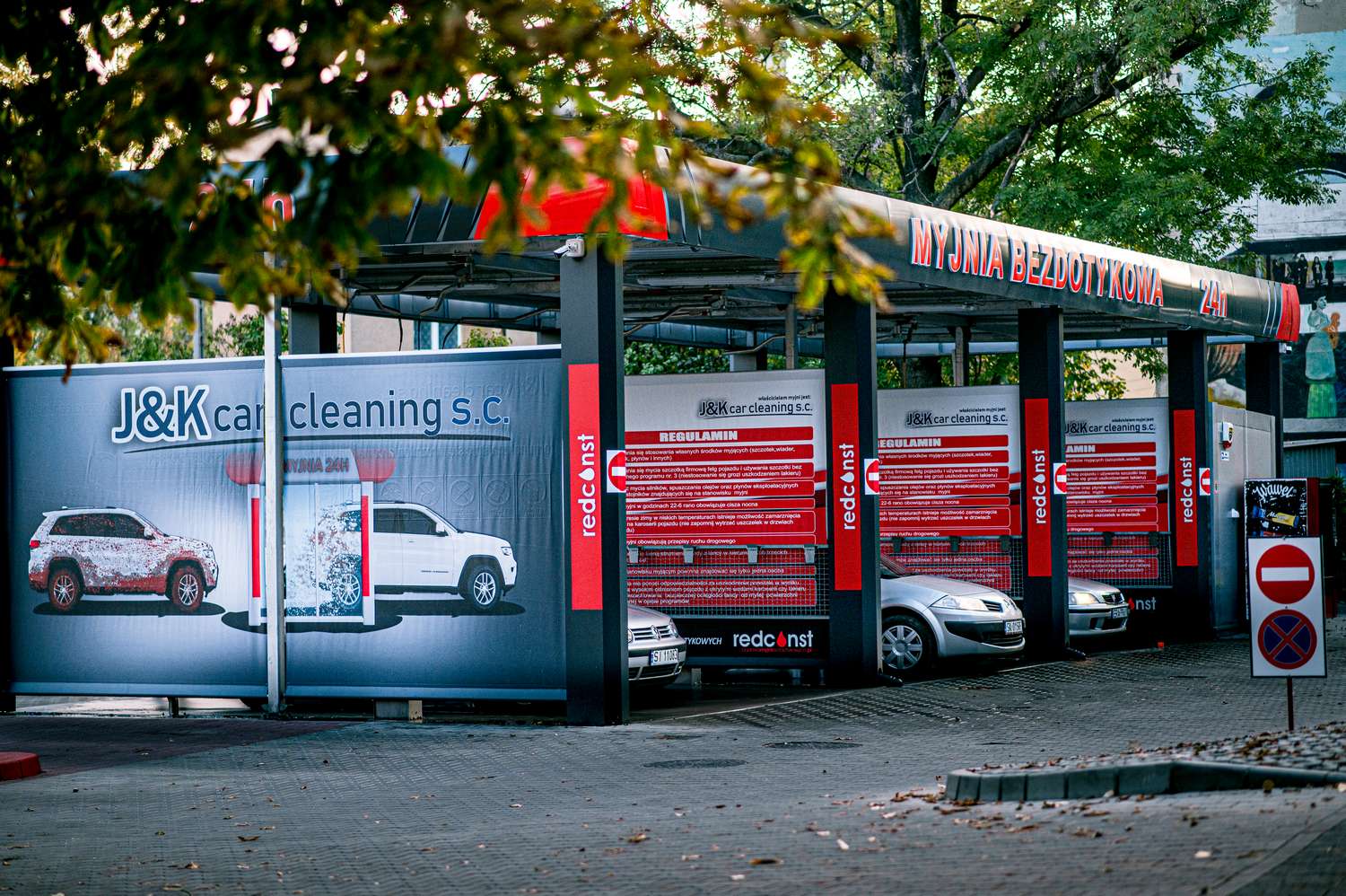 Myjnia bezdotykowa J&K car cleaning, oferująca nowoczesne usługi czyszczenia samochodów. Widoczne pojazdy i reklamy z informacjami o dostępnych programach myjni. Przestrzeń zaprojektowana dla wygodnego i efektywnego mycia aut, dostępna 24/7.