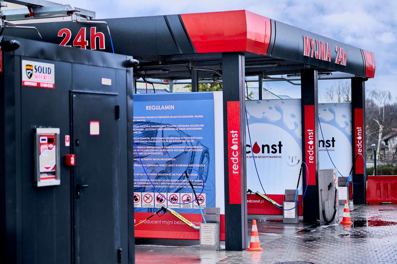 Widok myjni bezdotykowej działającej 24 godziny na dobę, z wyraźnym regulaminem i zabezpieczeniami. Elementy takie jak wąż do mycia oraz oznaczenia dotyczące zasad użytkowania podkreślają nowoczesność i komfort korzystania z usług producenta myjni bezdotykowych Redconst.