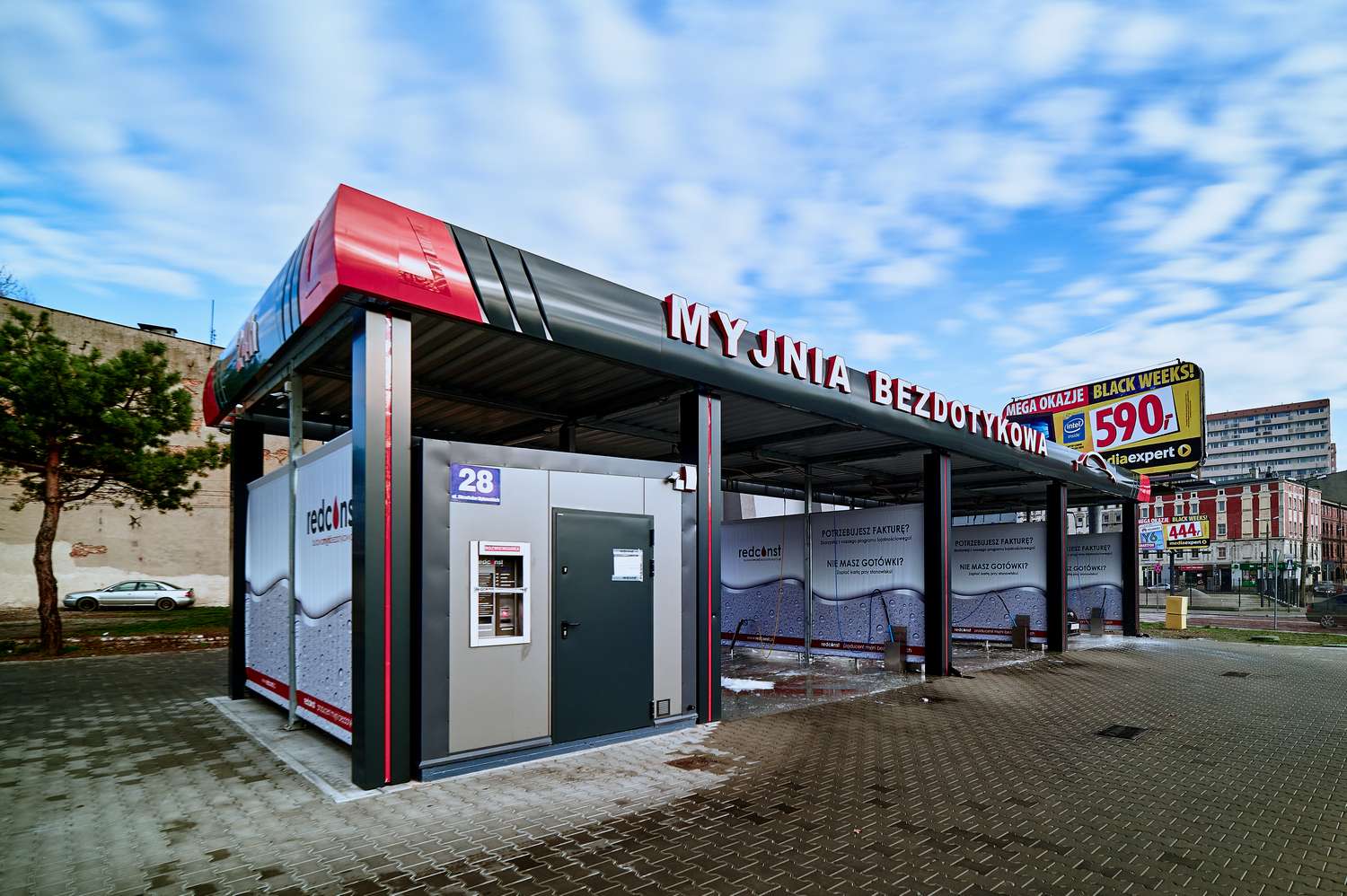 Obiekt myjni bezdotykowej z numerem 28, z nowoczesną konstrukcją i reklamą promocyjną, otoczony placem z betonową kostką. W tle widoczny krajobraz miasta i drzewo, co nadaje przestrzeni przyjemny charakter. Idealne miejsce do efektywnego mycia pojazdów.