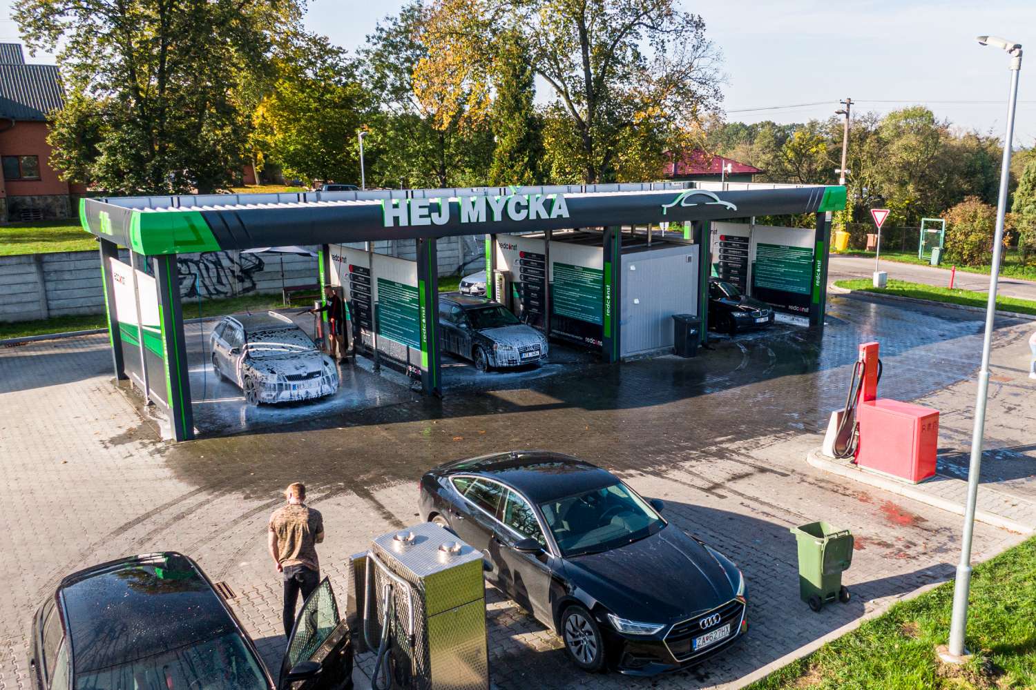 Widok na nowoczesną myjnię bezdotykową z kilkoma samochodami, które są myte. W tle widać drzewa oraz inne pojazdy. Myjnia oferuje różnorodne usługi czyszczenia pojazdów, a na jej elewacji znajdują się informacje o dostępnych opcjach.