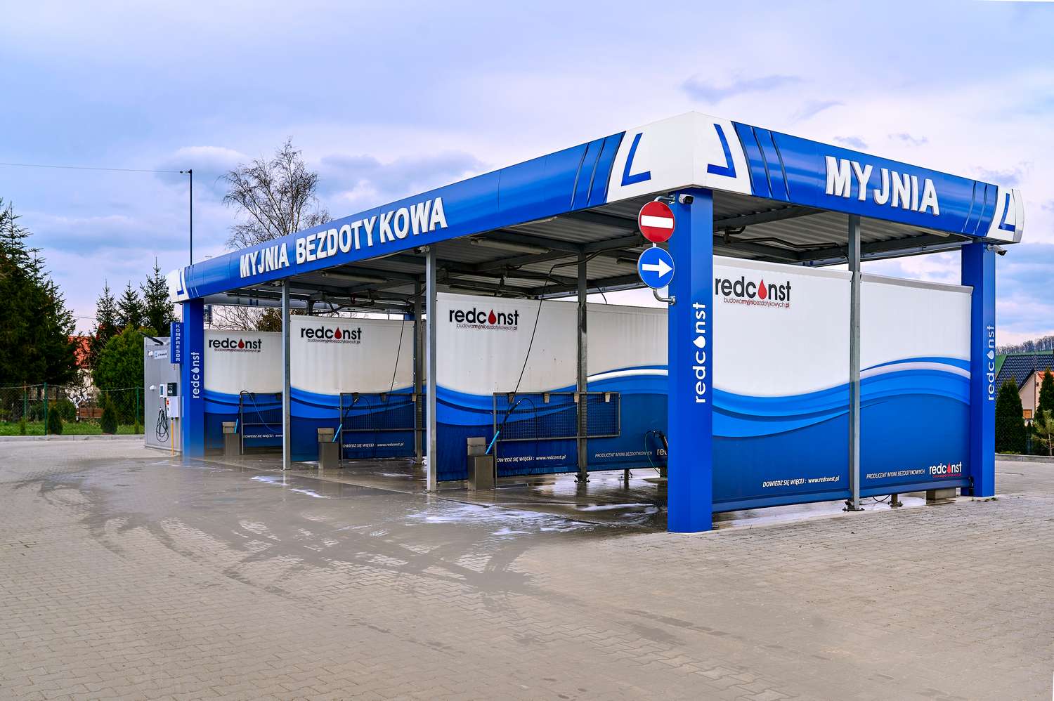 Stacja myjni bezdotykowej z nowoczesną konstrukcją, z widocznymi stanowiskami mycia samochodów. Na tle błękitnego nieba i zieleni, znak informujący o myjni oraz logo producenta. Przygotowana do użycia, z mokrą nawierzchnią po myciu. Idealne miejsce dla właścicieli aut szukających skutecznego czyszczenia.