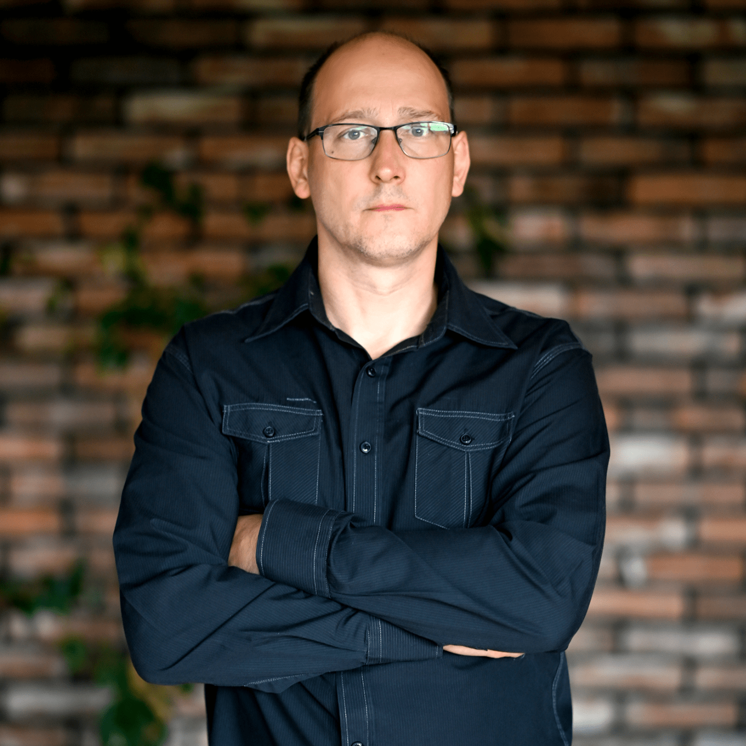 Mężczyzna w okularach, ubrany w czarną koszulę z długim rękawem, stoi z założonymi rękami. Tło stanowi ceglana ściana z roślinnością, co nadaje przytulny klimat. Jego poważny wyraz twarzy wskazuje na determinację i profesjonalizm, idealny dla producenta myjni bezdotykowej.