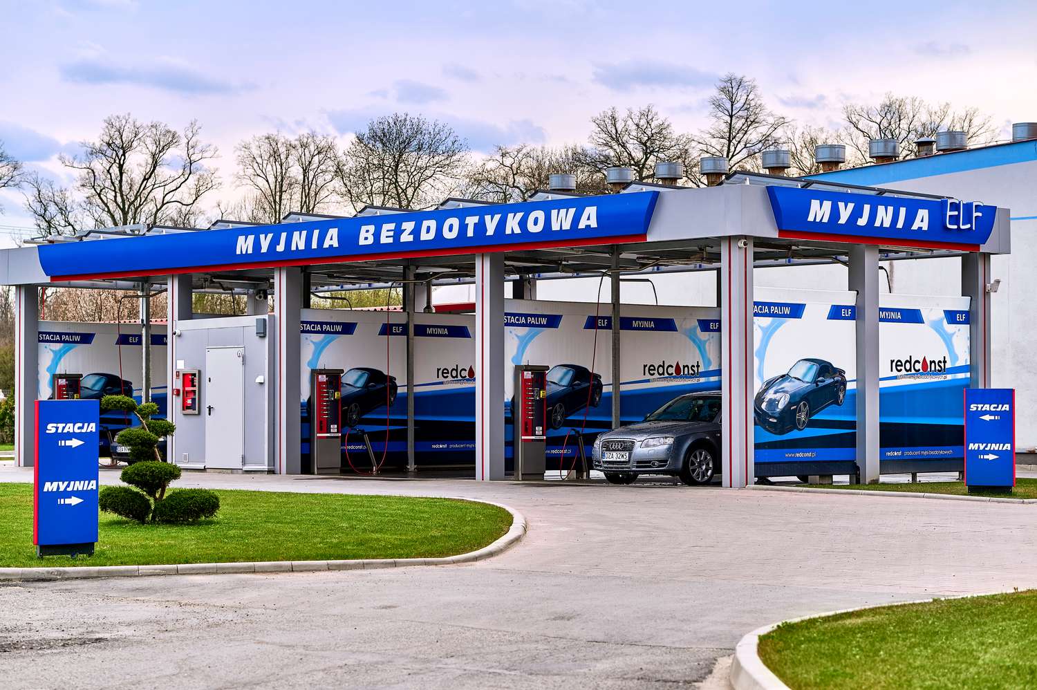 Obiekt myjni bezdotykowej z logo ELF, z przestrzenią do mycia samochodów. Widać kilka stanowisk, z nowoczesnym wyposażeniem i samochodami. Zielona trawa i ozdobne krzewy w tle. Idealne miejsce dla właścicieli pojazdów szukających łatwego i skutecznego sposobu na czyszczenie auta.
