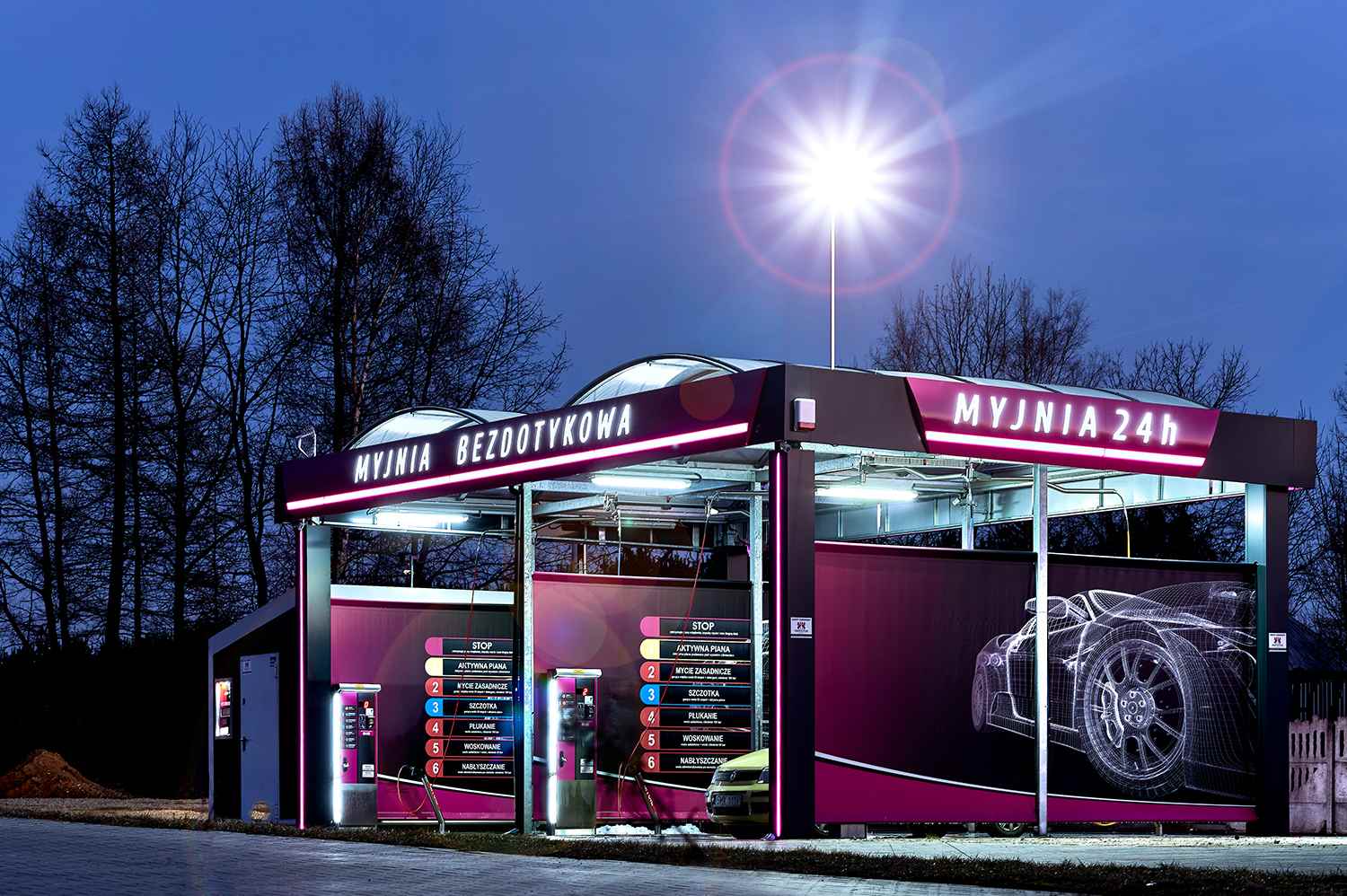 Zewnętrzny widok nowoczesnej myjni bezdotykowej, otwartej 24 godziny na dobę, z jasnym oświetleniem i informacyjnymi panelami dotyczącymi dostępnych usług. W tle ciemne niebo i nagie drzewa, a na pierwszym planie stacja myjni z widocznym samochodem.