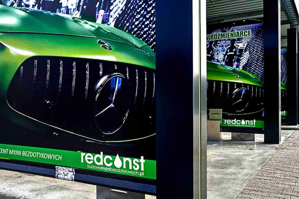 Reklama przedstawiająca zielony samochód marki Mercedes, z widocznym logo oraz hasłem "Producent myjni bezdotykowych". Tło z elementami miejskiego krajobrazu, podkreślające nowoczesność myjni. Idealne do promocji myjni bezdotykowych.