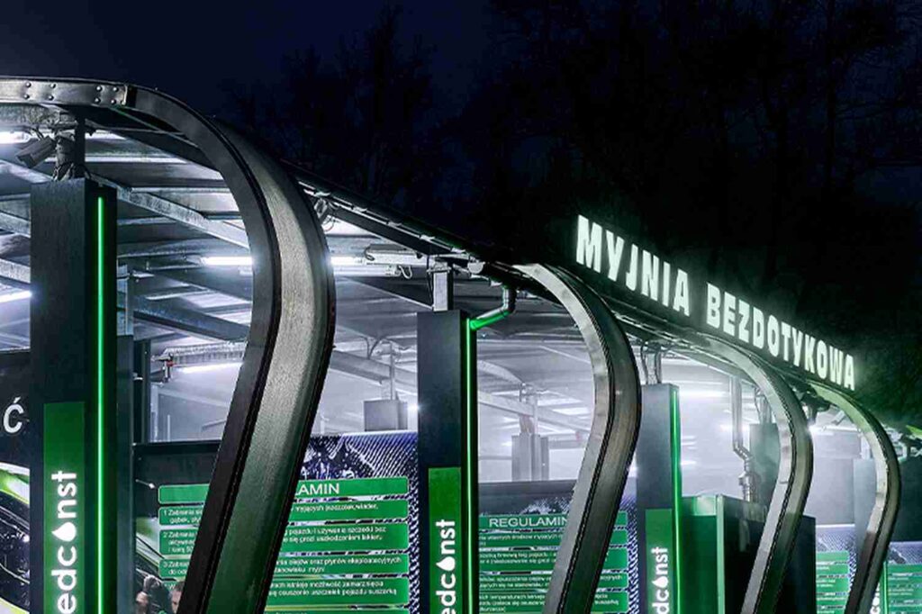 Nowoczesna myjnia bezdotykowa nocą, z podświetlonym napisem "MYJNIA BEZDOTYKOWA" i zielonymi detalami, podkreślającymi technologię i innowacyjność. W tle widoczne są instrukcje użytkowania oraz elegancka architektura obiektu, idealna dla miłośników czystych samochodów.