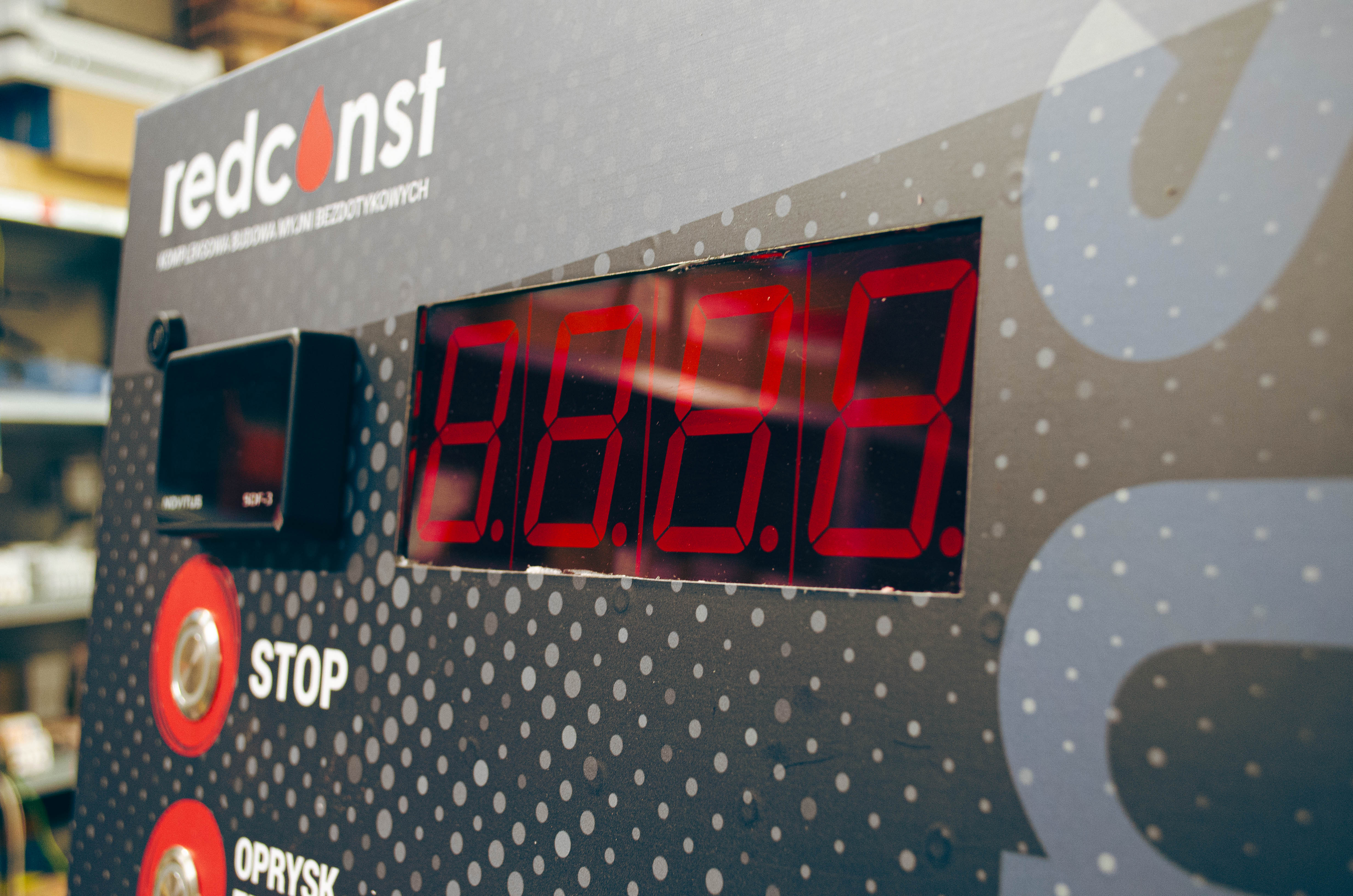 Panel sterujący myjni bezdotykowej z wyświetlaczem LED, na którym widoczne są cyfry 0.0.0 oraz przyciski "STOP" i "OPRYSK". Producent myjni bezdotykowej Redconst, nowoczesne rozwiązania dla czystości samochodów.