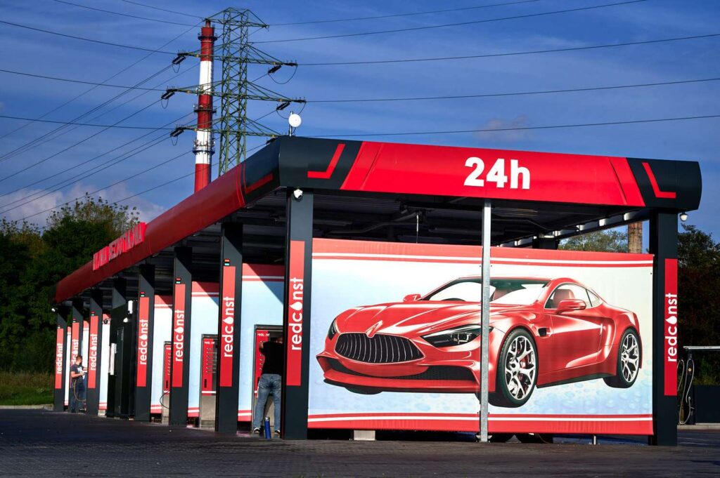 Nowoczesna myjnia bezdotykowa otwarta 24 godziny na dobę, z czerwonymi akcentami i dużą reklamą eleganckiego samochodu. W tle widoczne są słupy energetyczne, co podkreśla przemysłowy charakter lokalizacji. Idealne miejsce dla właścicieli pojazdów szukających wygodnych usług mycia.